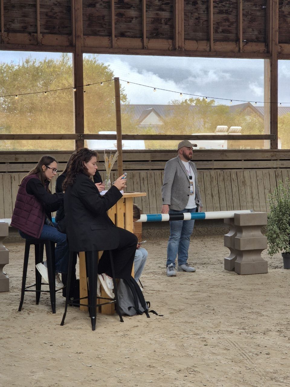 Visitors working at a standing table on the sand of the covered arena, a show jump pole resting on its blocks behind them