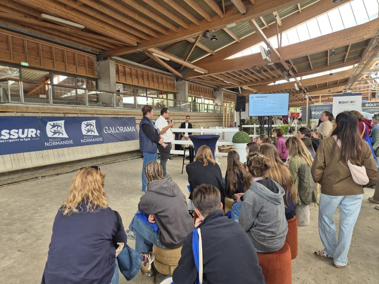 A talk in progress in the arena, two speakers sharing a microphone in front of the slides while the audience sits on stools on the sand