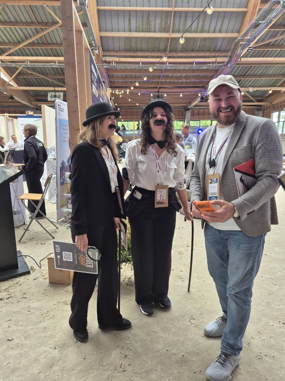 Three visitors laughing over a phone between the stands, two of them in bowler hats and false moustaches for the event's quiz