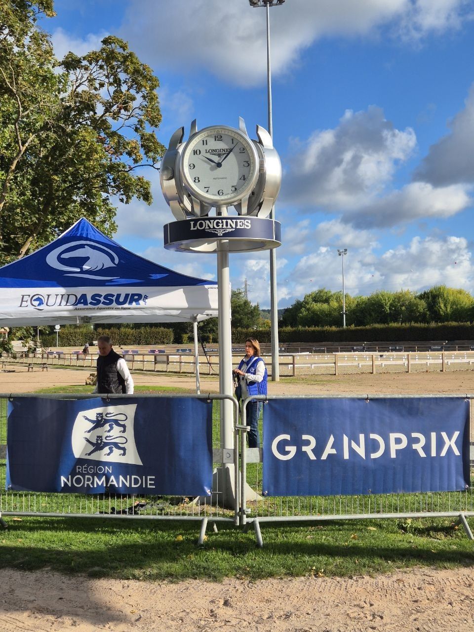 The Longines clock on its post beside the outdoor arena, Région Normandie and GrandPrix banners on the fence below it