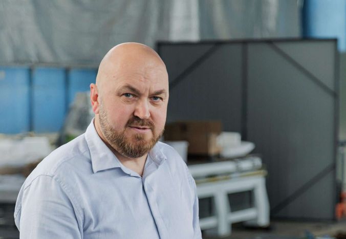 EHOSS founder Viktor Kvachan in the production hall, looking into the camera, a braced steel panel and a white machine frame out of focus behind him