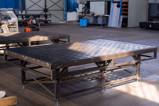 Two EHOSS welding tables standing in the hall, the nearer one's slotted steel top on its braced frame and levelling feet